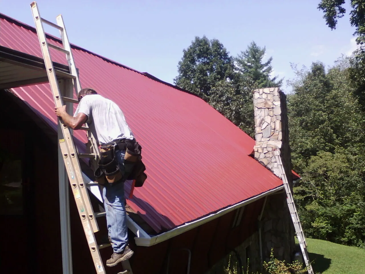 Roofer on a ladder beside a red metal roof and stone chimney — Soffit & Fascia Repair by Roofing Brothers in Mc Connells
